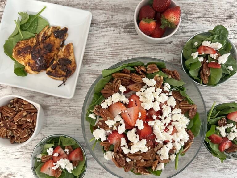 Large bowl of spinach strawberry salad with smaller bowls of salad, a plate of cooked chicken breast, bowl of strawberries, and bowl of pecans.