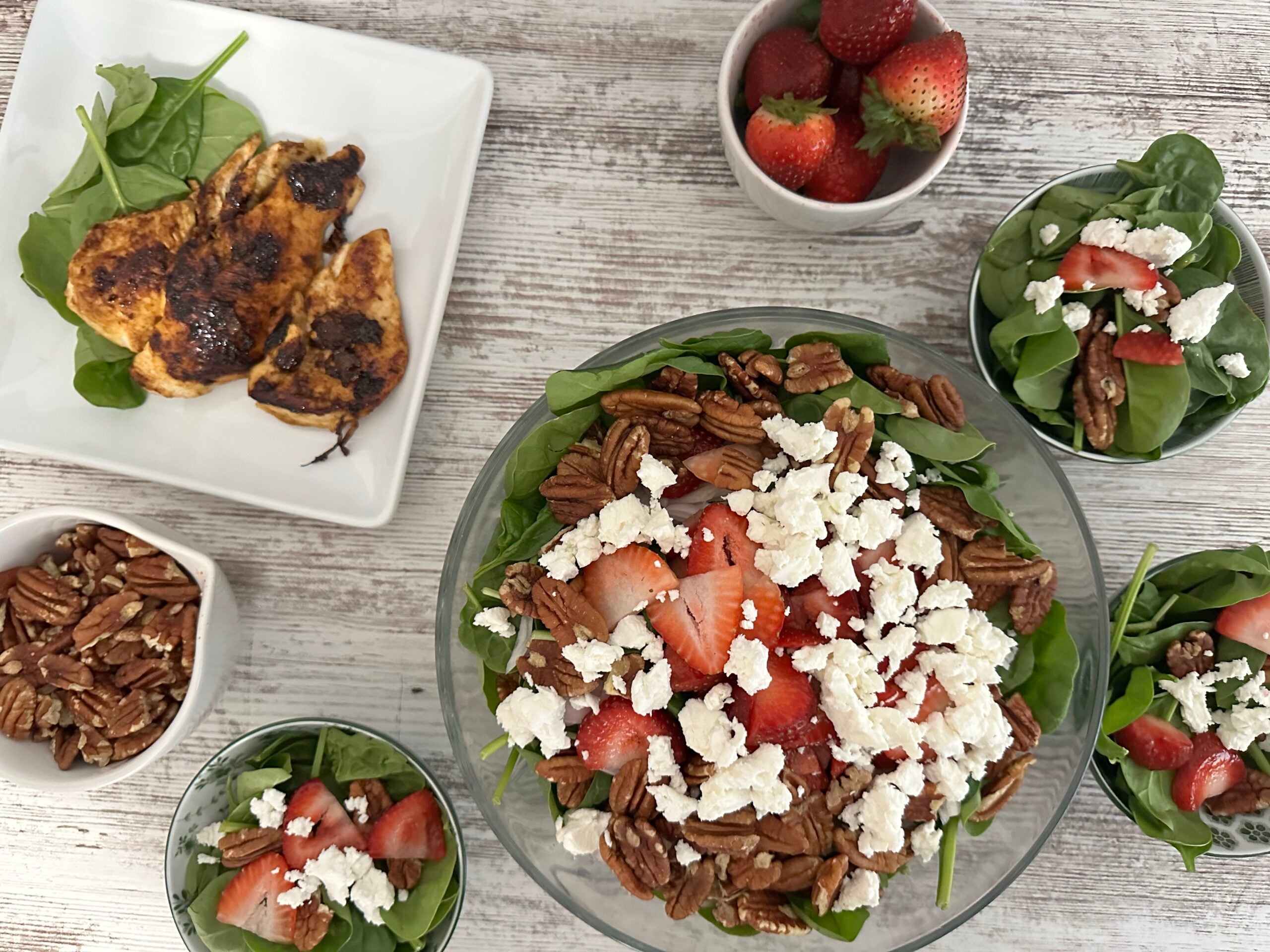 Large bowl of spinach strawberry salad with smaller bowls of salad, a plate of cooked chicken breast, bowl of strawberries, and bowl of pecans.