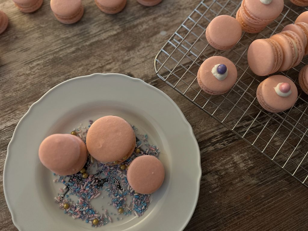 Four vanilla rose macarons on a dish with colorful sprinkles. Macarons on a baking rack toward the top.