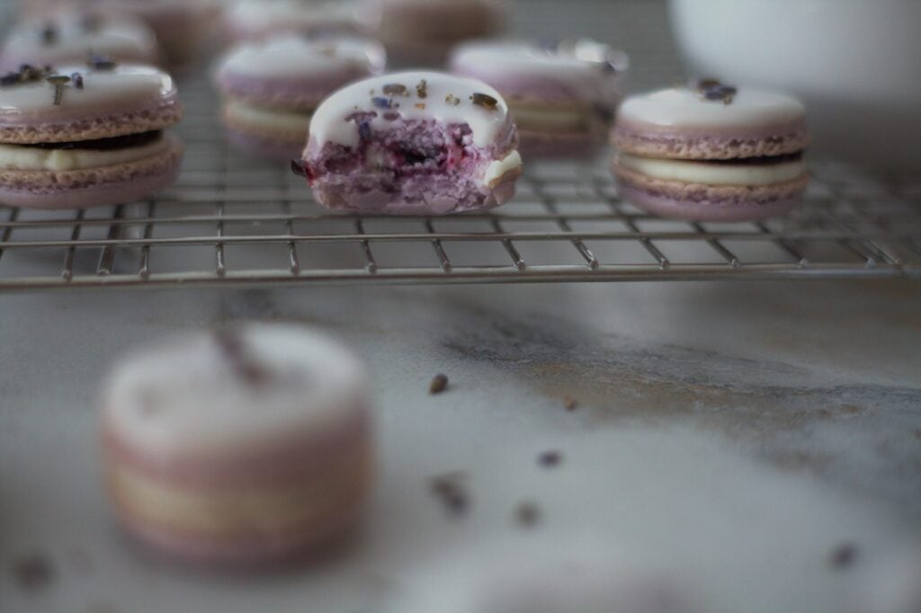 Blueberry lavender French macarons on a baking rack, one with a bite taken out of it.
