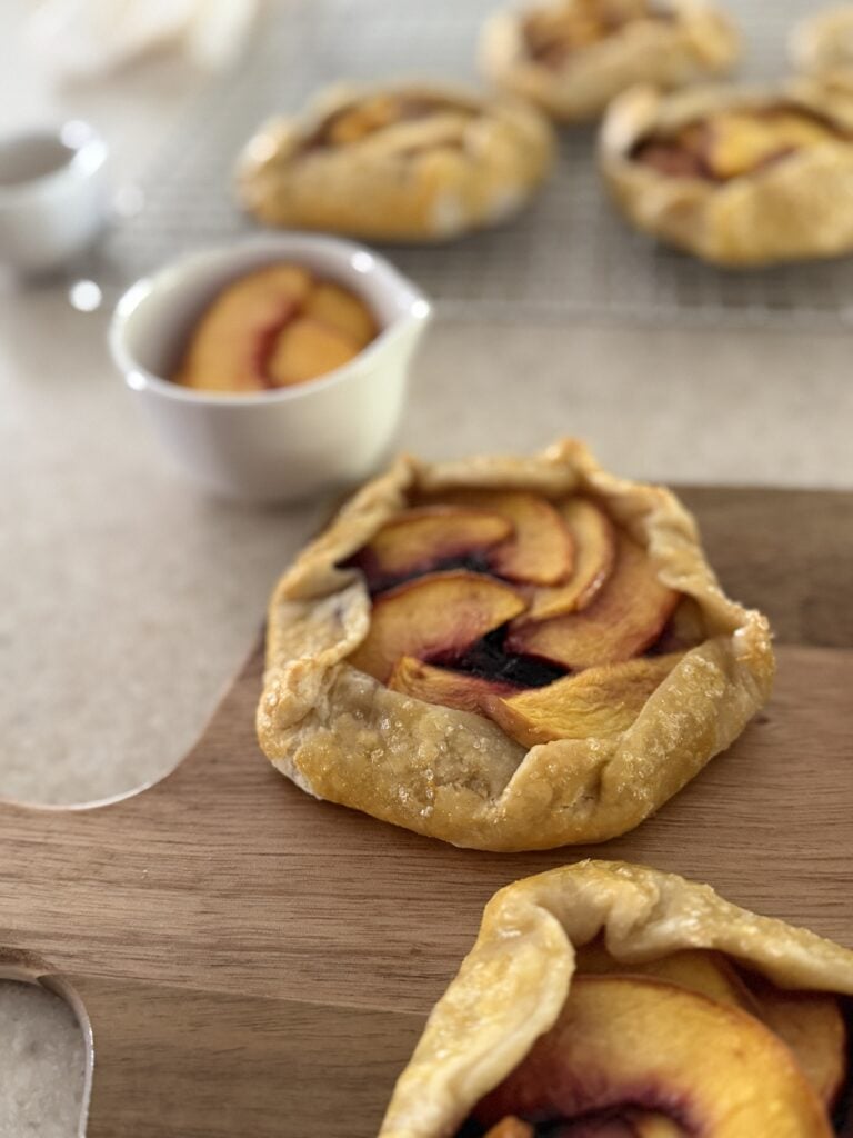 Two peach blueberry mini galettes on a cutting board with sliced peaches and more mini galettes in the background.