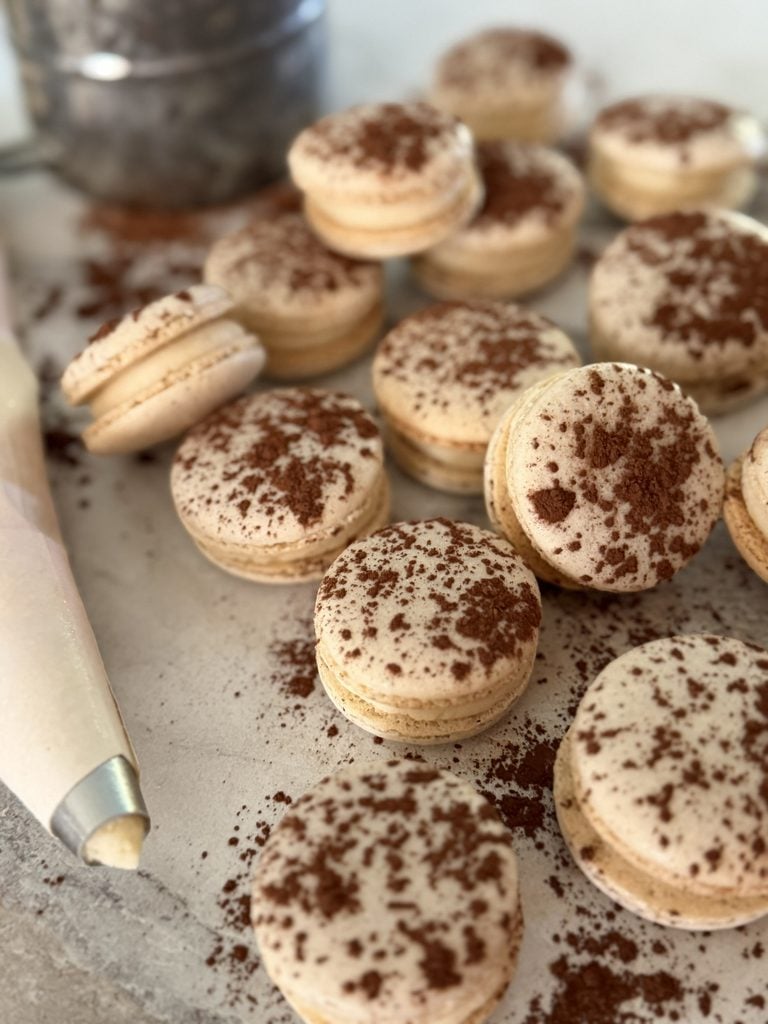 Tiramisu macarons arranged on a surface with a cup in the background and a piping on the left.